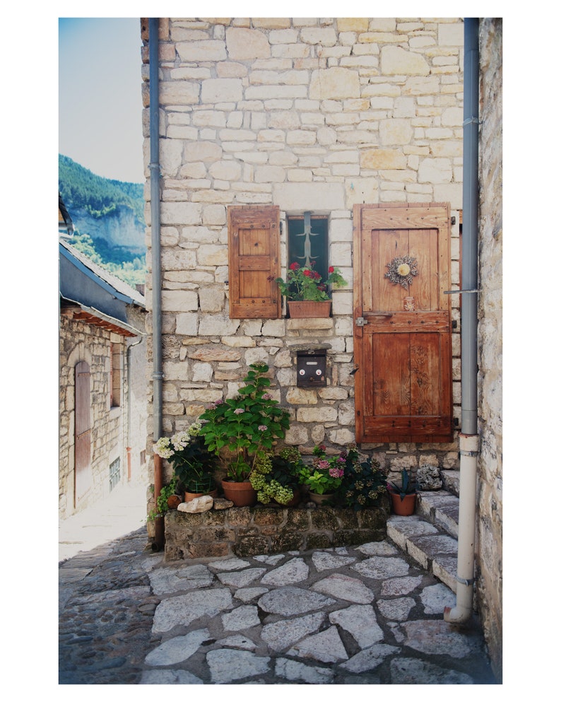 May include: A stone building with a wooden door and a window with shutters. There are potted plants in front of the building and a stone path.