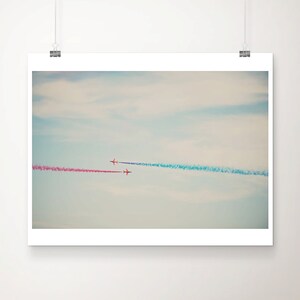 May include: A framed print featuring two red airplanes leaving trails of red and blue smoke against a pale blue sky. The artwork is mounted with silver clips, suggesting a focus on aviation and aerial displays.