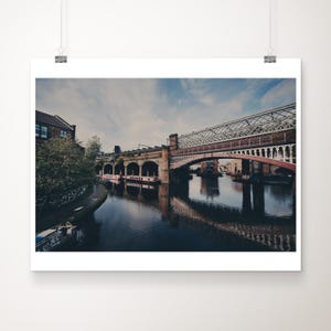 May include: A view of a canal with a brick bridge and a metal bridge over it. The water is calm and reflects the bridges and the buildings on the shore.