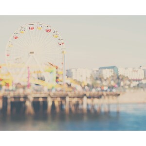 May include: A soft-focus photograph of a Ferris wheel with red, yellow, and white gondolas. The wheel is set against a pale sky and a blurred background of buildings and a pier. The water in the foreground is a calm blue.