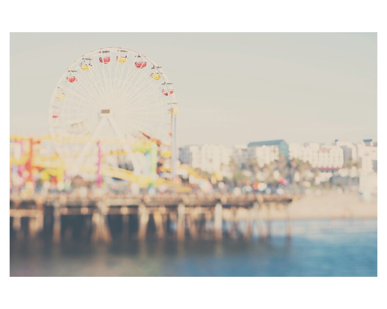 May include: A soft-focus photograph of a Ferris wheel with red, yellow, and white gondolas. The wheel is set against a pale sky and a blurred background of buildings and a pier. The water in the foreground is a calm blue.