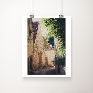 May include: A narrow, cobblestone street in a European village with stone buildings on either side. The buildings are old and have a rustic charm. The street is lined with lush greenery and the sun is shining brightly.