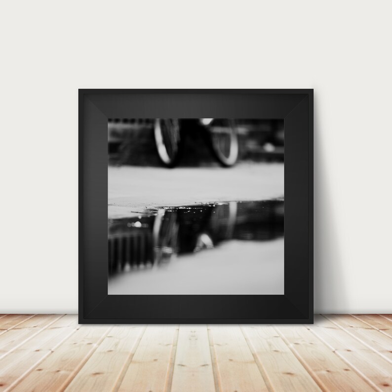 May include: Black and white photograph of a bicycle reflected in a puddle. The image is framed in a black square frame, mounted on a white wall above a wooden floor.