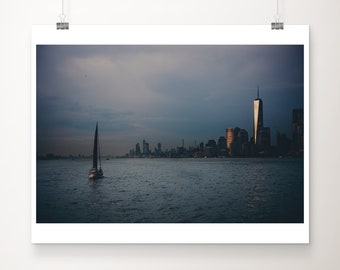 May include: A sailboat sails towards the New York City skyline at sunset. The city is silhouetted against a cloudy sky, with the Freedom Tower standing tall in the center.