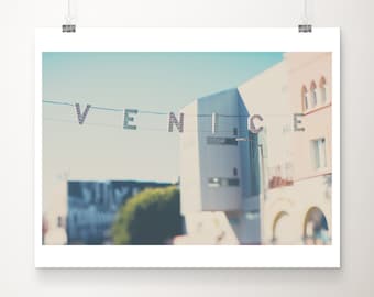 May include: A photograph of a building with the word "VENICE" strung across the top. The letters are attached to a wire and are in front of a blue sky. The buildings are light-colored and have a vintage aesthetic.
