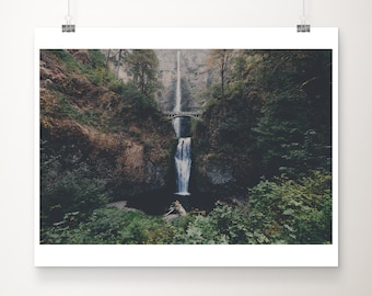 Può includere: Una cascata scende da una scogliera rocciosa, circondata da alberi verdi lussureggianti. Un piccolo ponte attraversa l'acqua in cima alla cascata.