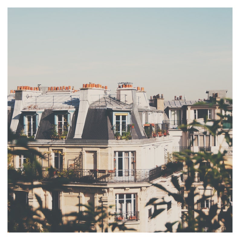 May include: A view of a Parisian apartment building with multiple windows and balconies. The building is painted in a light beige color with a dark gray roof. The building is surrounded by lush green foliage.