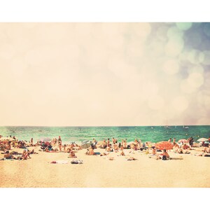 May include: A sunny beach scene with a large group of people enjoying the warm weather. The beach is sandy and the water is a bright turquoise blue. There are several umbrellas and beach chairs scattered throughout the scene.