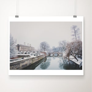 May include: A snowy winter scene with a bridge over a canal. The canal is lined with trees and a building in the background. The sky is a light gray.