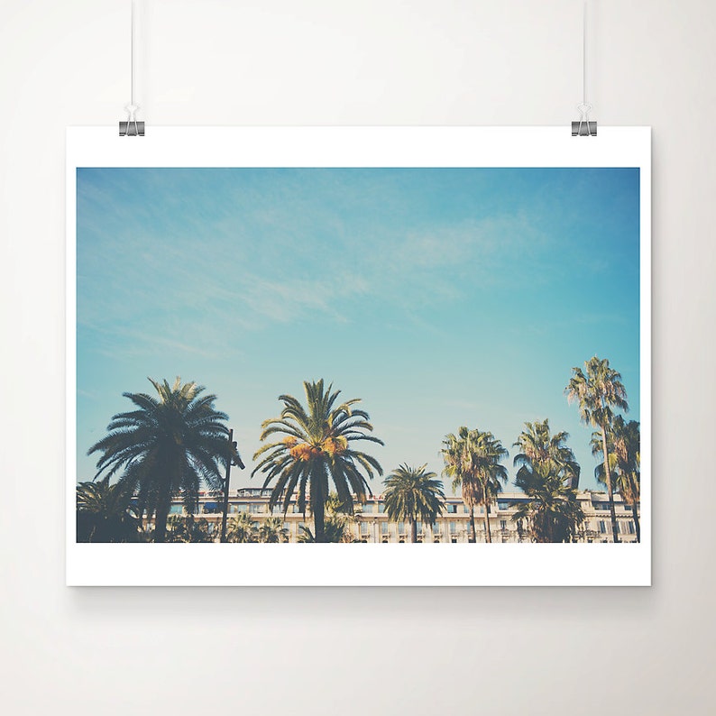 May include: A framed print showing palm trees under a clear blue sky, with a building in the background. The image has a vintage feel. The print is attached to a white wall with a silver clip. The image has a slightly faded aesthetic.