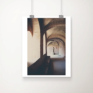 May include: A framed print depicting an arched hallway with brick and stone walls. The hallway features a series of arched ceilings and windows, with sunlight illuminating the space. The print is displayed against a white backdrop.