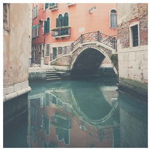 K&ouml;nnte beinhalten: Eine malerische Kanalszene in Venedig, Italien, mit einer Steinbr&uuml;cke mit kunstvollen schmiedeeisernen Gel&auml;ndern. Geb&auml;ude mit roten und pfirsichfarbenen Fassaden s&auml;umen den Kanal, mit Spiegelungen im Wasser. Gr&uuml;ne Fensterl&auml;den und Blumenk&auml;sten setzen Farbakzente.