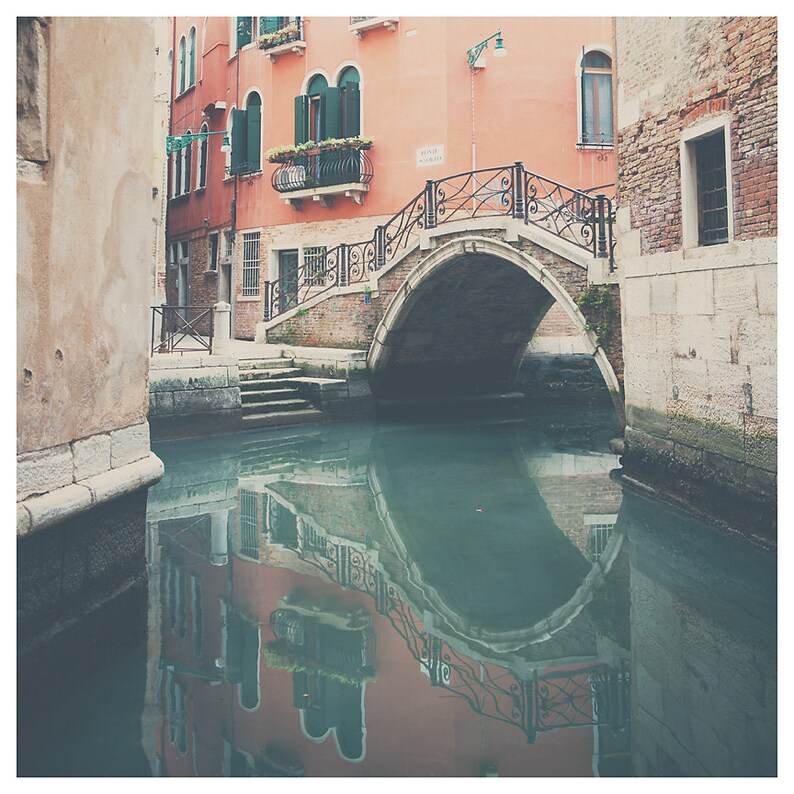 K&ouml;nnte beinhalten: Eine malerische Kanalszene in Venedig, Italien, mit einer Steinbr&uuml;cke mit kunstvollen schmiedeeisernen Gel&auml;ndern. Geb&auml;ude mit roten und pfirsichfarbenen Fassaden s&auml;umen den Kanal, mit Spiegelungen im Wasser. Gr&uuml;ne Fensterl&auml;den und Blumenk&auml;sten setzen Farbakzente.