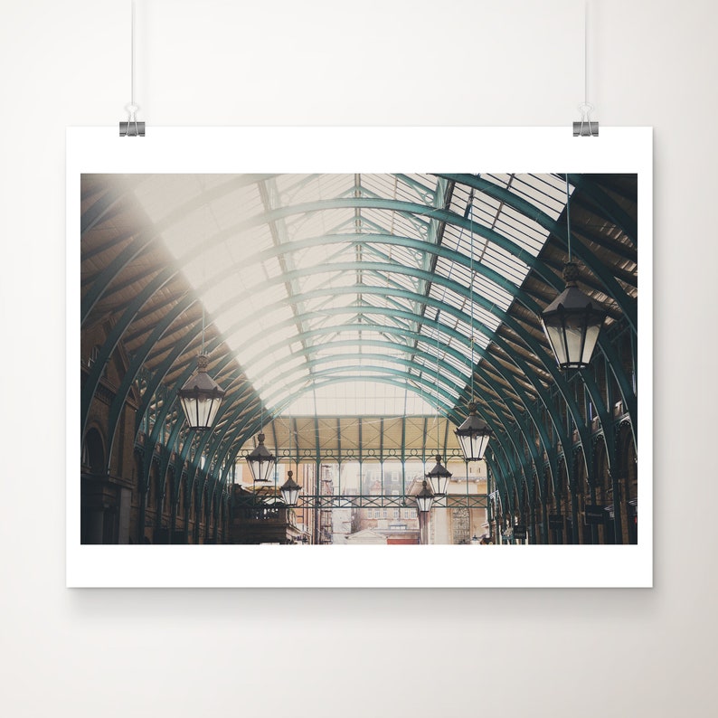 May include: A framed print depicting the interior of a historic building. The image shows a glass and metal arched ceiling with hanging lanterns. Sunlight enters from the left, lighting the space. The print is displayed on a white wall.