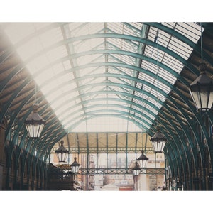 May include: An interior shot of a building with a curved glass and metal roof. Dark, ornate lanterns are suspended from the ceiling. Sunlight streams through the glass panels, lighting the space. The building's structure is visible in the background, with a view of the outside.