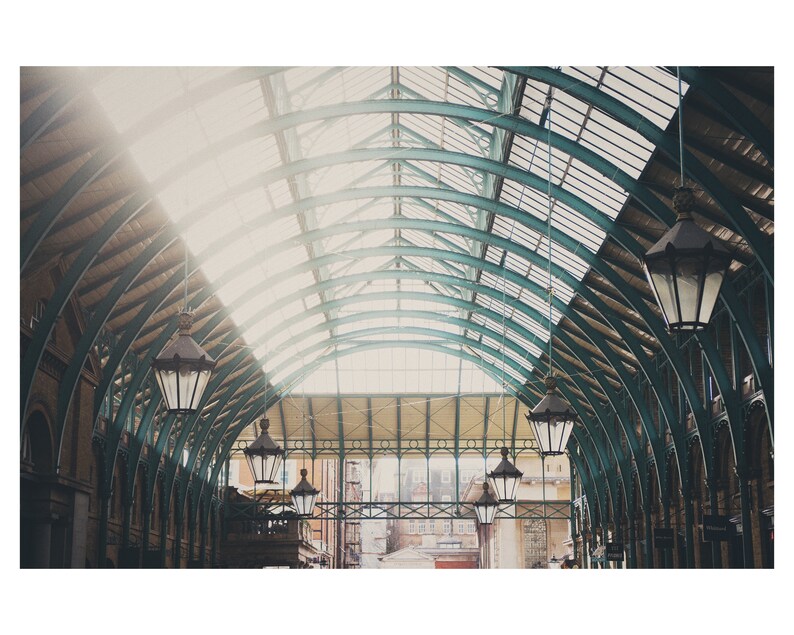 May include: An interior shot of a building with a curved glass and metal roof. Dark, ornate lanterns are suspended from the ceiling. Sunlight streams through the glass panels, lighting the space. The building's structure is visible in the background, with a view of the outside.
