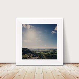 Puede incluir: Una fotografía enmarcada de un paisaje con un afloramiento rocoso en primer plano, campos verdes en el medio y un cielo azul con nubes blancas en el fondo.
