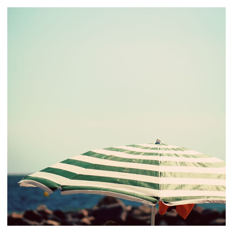 May include: A green and white striped beach umbrella with a wooden handle. The umbrella is open and is casting a shadow on the sand.