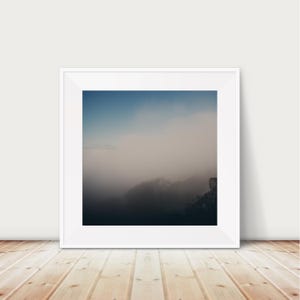 Puede incluir: Una fotografía enmarcada de un paisaje montañoso brumoso. El cielo es un degradado de azul y blanco, con algunas nubes tenues. Los picos de las montañas están ocultos por la niebla, creando una sensación de misterio y tranquilidad.