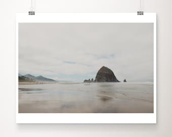 May include: A landscape print featuring a coastal scene with a large rock formation in the ocean. The beach is wet and reflective, with a cloudy sky overhead. The print is displayed on a white background.