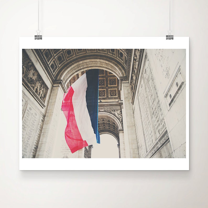 May include: A framed print featuring the Arc de Triomphe in Paris, France, with a French flag hanging in the center. The flag displays vertical stripes of red, white, and blue. The architecture is detailed with carvings and inscriptions.