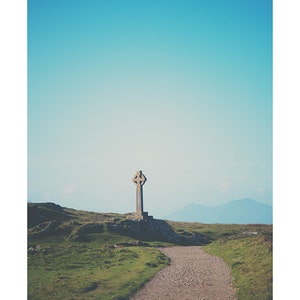 May include: A stone Celtic cross stands on a grassy hilltop with a dirt path leading towards it. The sky is a clear blue with distant mountains in the background.