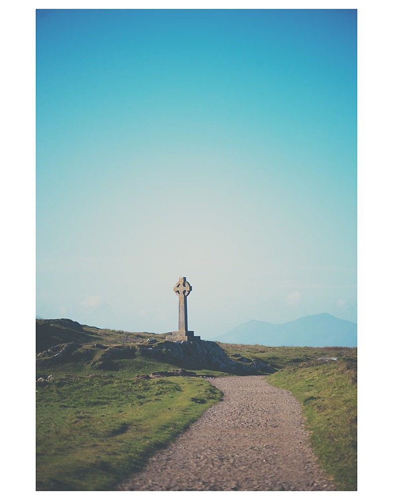 May include: A stone Celtic cross stands on a grassy hilltop with a dirt path leading towards it. The sky is a clear blue with distant mountains in the background.