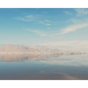 May include: A still, reflective body of water with a mountain range in the distance. The sky is a light blue with wispy white clouds.