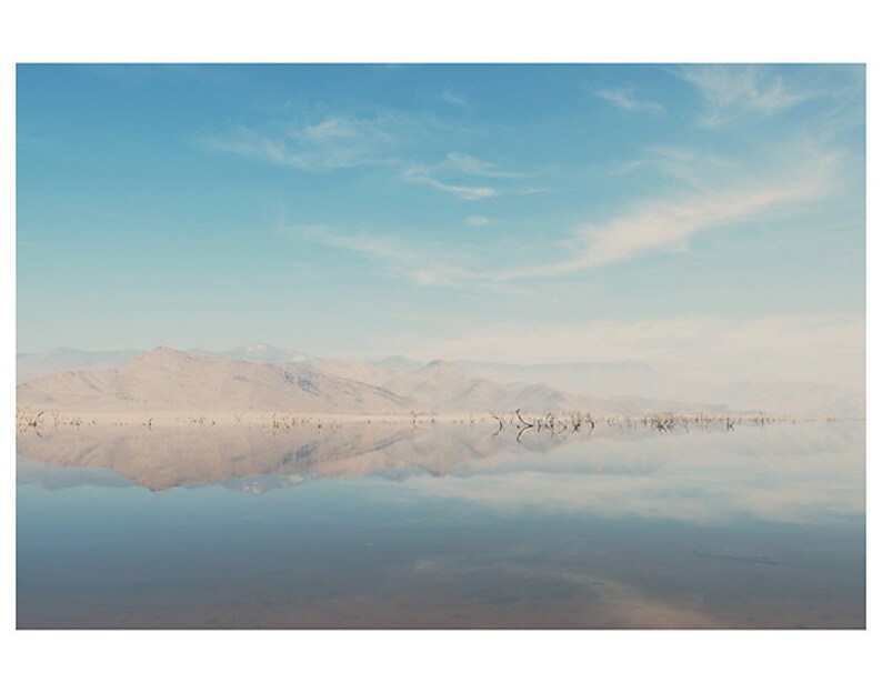 May include: A still, reflective body of water with a mountain range in the distance. The sky is a light blue with wispy white clouds.
