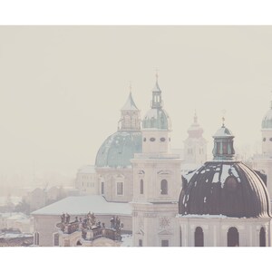 May include: A snowy cityscape with several church domes, including a large, dark green dome with a smaller, lighter green dome on top. The buildings are white and have a vintage, faded look.