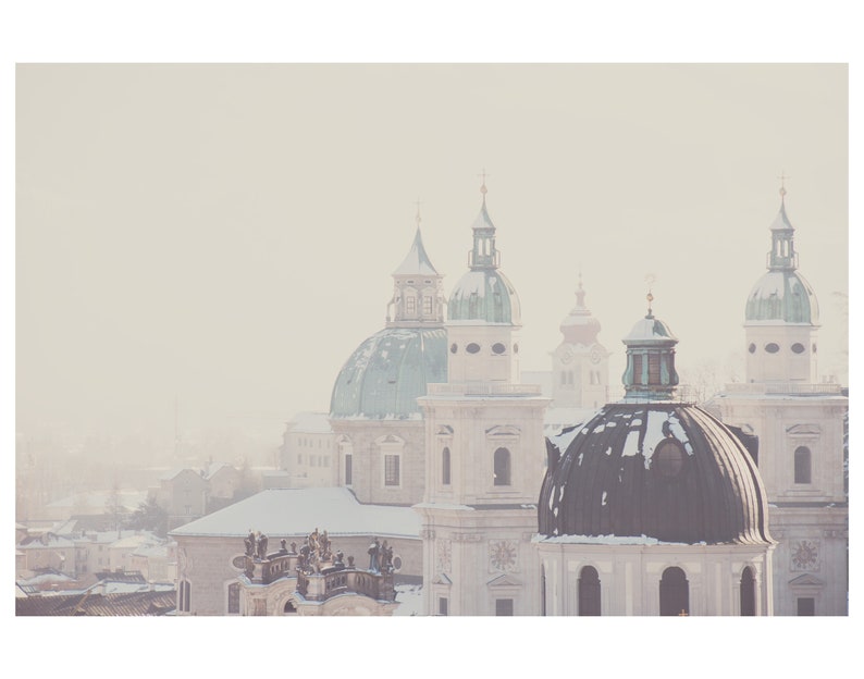 May include: A snowy cityscape with several church domes, including a large, dark green dome with a smaller, lighter green dome on top. The buildings are white and have a vintage, faded look.