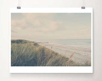 Op de afbeelding: Een serene strandscène-afdruk. De afbeelding toont een zandstrand, een kalme oceaan en een bewolkte lucht. Hoge, droge grassen omzomen de voorgrond. De afdruk wordt op een witte achtergrond weergegeven, vastgehouden door zilveren clips.