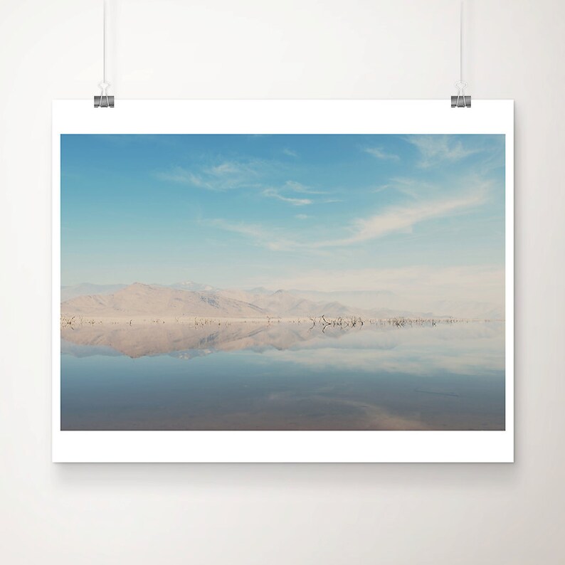 May include: A photograph of a still, reflective body of water with a mountain range in the background. The sky is a light blue with white clouds.
