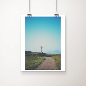 May include: A stone Celtic cross stands on a grassy hilltop with a dirt path leading towards it. The sky is a bright blue with a few white clouds.