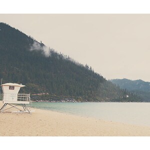 May include: A serene beach scene with a white lifeguard tower on the sand. The turquoise water meets the sandy shore, with a backdrop of a forested mountain under a cloudy sky. The image evokes a sense of calm and natural beauty.