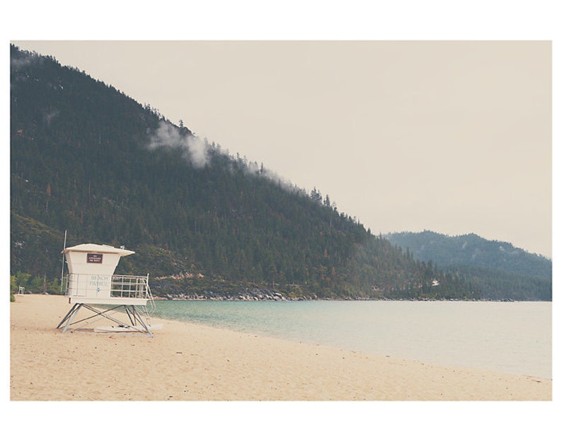 May include: A serene beach scene with a white lifeguard tower on the sand. The turquoise water meets the sandy shore, with a backdrop of a forested mountain under a cloudy sky. The image evokes a sense of calm and natural beauty.