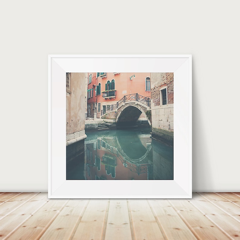 K&ouml;nnte beinhalten: Ein gerahmter Druck einer venezianischen Kanalszene. Das Bild zeigt eine Steinbr&uuml;cke, die sich &uuml;ber dem Wasser w&ouml;lbt, mit Geb&auml;uden in Pfirsich- und Beiget&ouml;nen, die den Kanal s&auml;umen. Das Wasser spiegelt die Br&uuml;cke und die Geb&auml;ude wider. Der Druck ist in einem wei&szlig;en Rahmen.