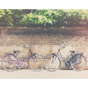Op de afbeelding: Twee vintage fietsen met rieten manden staan tegen een bakstenen muur. E&eacute;n fiets is paars, de andere zwart. Groen gebladerte bedekt de muur en cre&euml;ert een klassieke, nostalgische sc&egrave;ne.