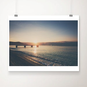 May include: A framed print of a serene beach scene at sunrise. The image captures a pier extending into calm water, with the sun rising over the horizon. The sky transitions from deep blue to golden hues, reflecting on the water.
