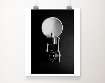 Op de afbeelding: Zwart-wit foto van een vintage camera met een flitser. De camera staat centraal, met een grote, ronde flitsreflector erboven. De foto is op papier gedrukt en opgehangen met klemmen.