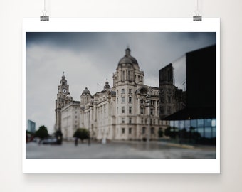 May include: A framed print of a large, ornate building with multiple domes and a clock tower. The building is light-colored, possibly stone, and set against a cloudy sky. A modern, dark building is on the right.