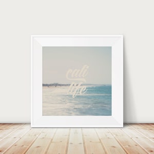 Puede incluir: Una impresi&oacute;n enmarcada que presenta una escena de playa con las palabras "cali life" en una fuente cursiva. La imagen muestra una playa arenosa, olas del oc&eacute;ano y un cielo azul claro. El marco es blanco.