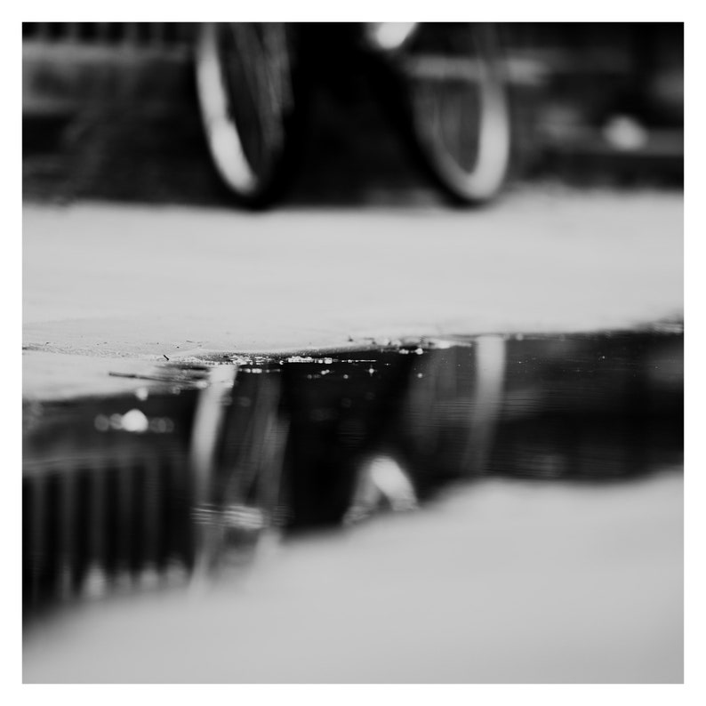 May include: Black and white photograph of a bicycle with a reflection in a puddle. The bicycle wheels are blurred in the background, while the reflection in the water is in sharp focus. The image is taken outdoors.