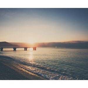 May include: A serene seascape at sunrise. The image captures a concrete pier extending into calm, turquoise water. The sun, a bright orb, is positioned at the horizon, casting a warm glow over the scene. The sky transitions from a deep blue to a soft, golden hue.