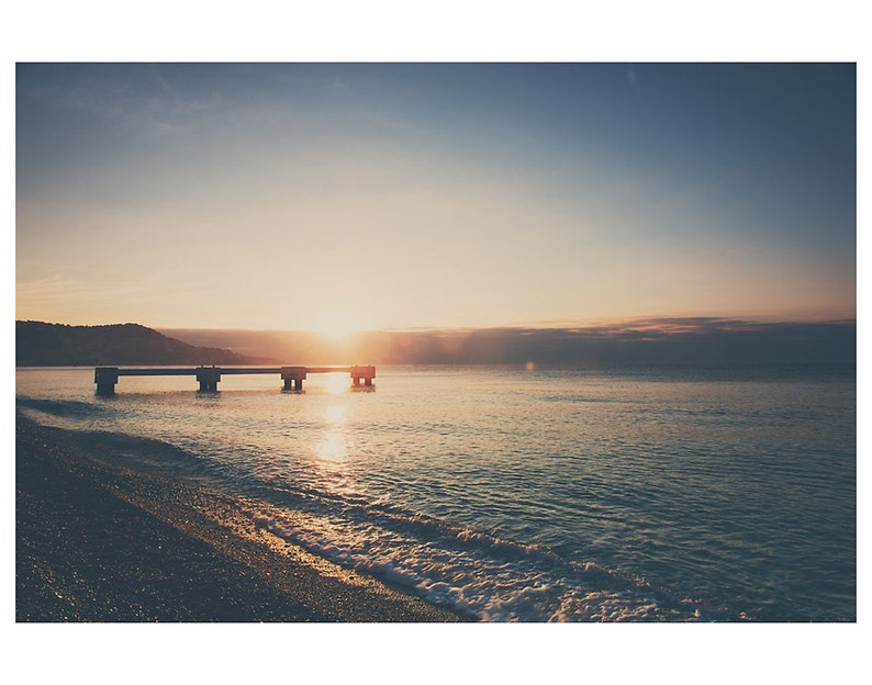 May include: A serene seascape at sunrise. The image captures a concrete pier extending into calm, turquoise water. The sun, a bright orb, is positioned at the horizon, casting a warm glow over the scene. The sky transitions from a deep blue to a soft, golden hue.