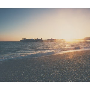 May include: A scenic coastal view at sunset. The image features a pier extending into the ocean, with the sun setting in the background. The foreground shows a pebble beach and the ocean waves.