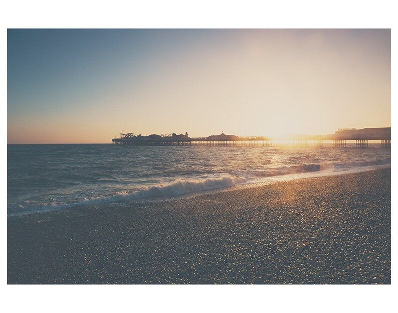 May include: A scenic coastal view at sunset. The image features a pier extending into the ocean, with the sun setting in the background. The foreground shows a pebble beach and the ocean waves.
