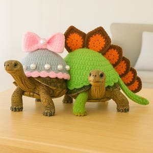 Op de afbeelding: Twee decoratieve schildpadbeeldjes. Eén schildpad draagt een grijze gehaakte hoed met een roze strik en parelaccenten. De andere schildpad heeft een groene gehaakte schild met bruine en oranje accenten.