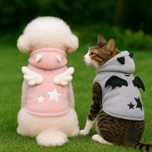 May include: A white dog and a tabby cat wearing matching hooded vests. The dog's vest is pink with white angel wings and star accents. The cat's vest is gray with black bat wings and star accents. Both are sitting on green grass.