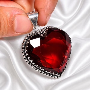 May include: A heart-shaped pendant with a large, faceted, deep red gemstone. The gemstone is set in a silver-colored metal frame with a beaded border and a triangular bail for a chain. The pendant is held against a white, silky background.
